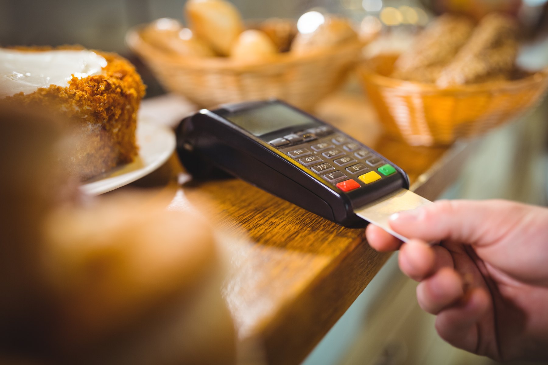 A hand inserting a card into a black payment terminal on a wooden bakery counter, with baskets of bread and a cake blurred in the background.