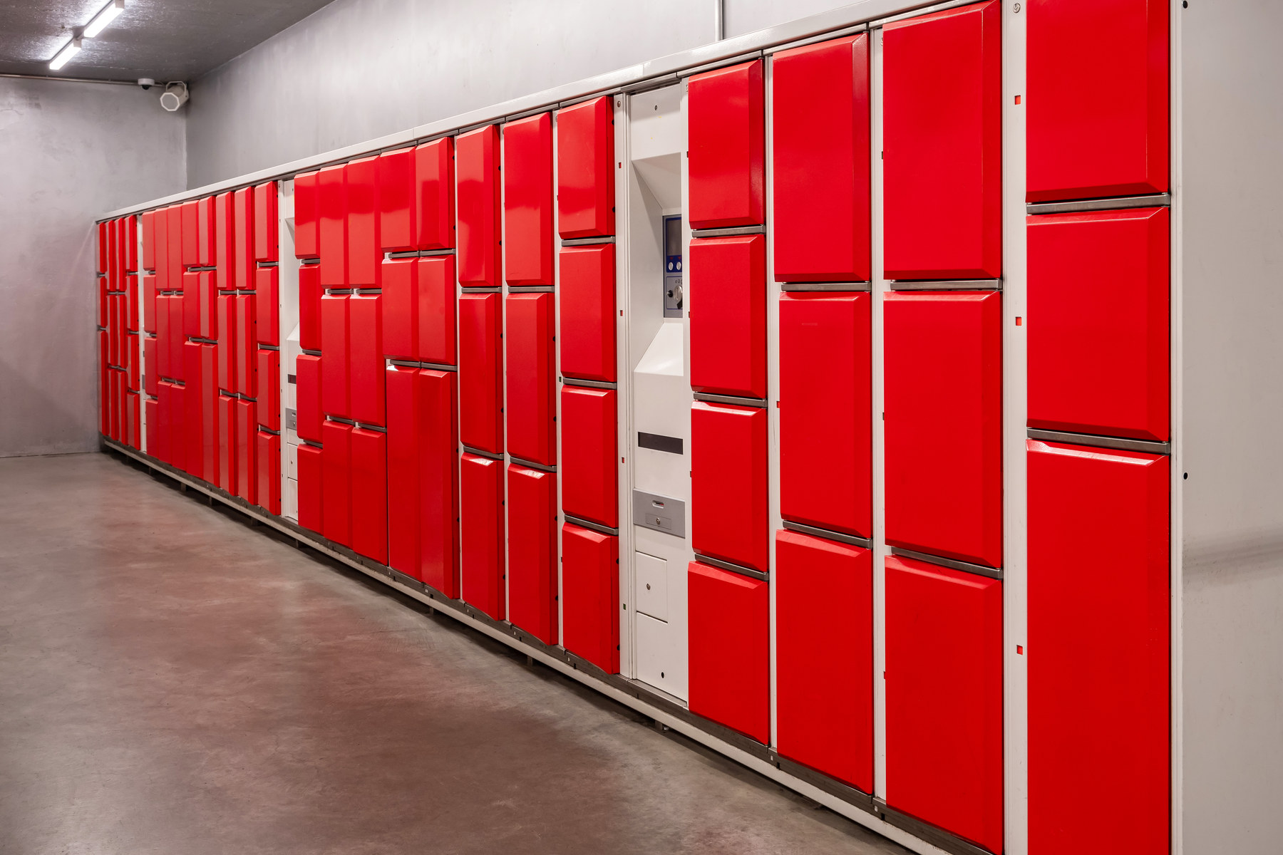 A long bank of bright red luggage lockers of varied sizes set in white metal frames against a bare concrete wall.