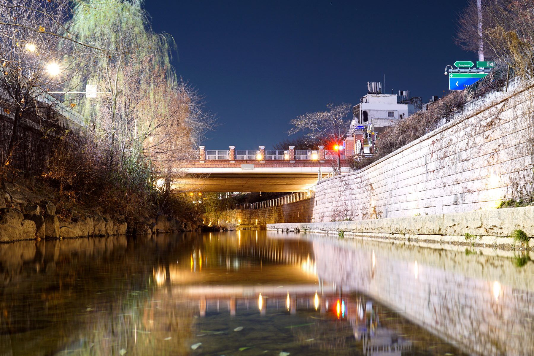 Cheonggyecheon stream at night, lamplight reflected in the water beneath a road bridge.