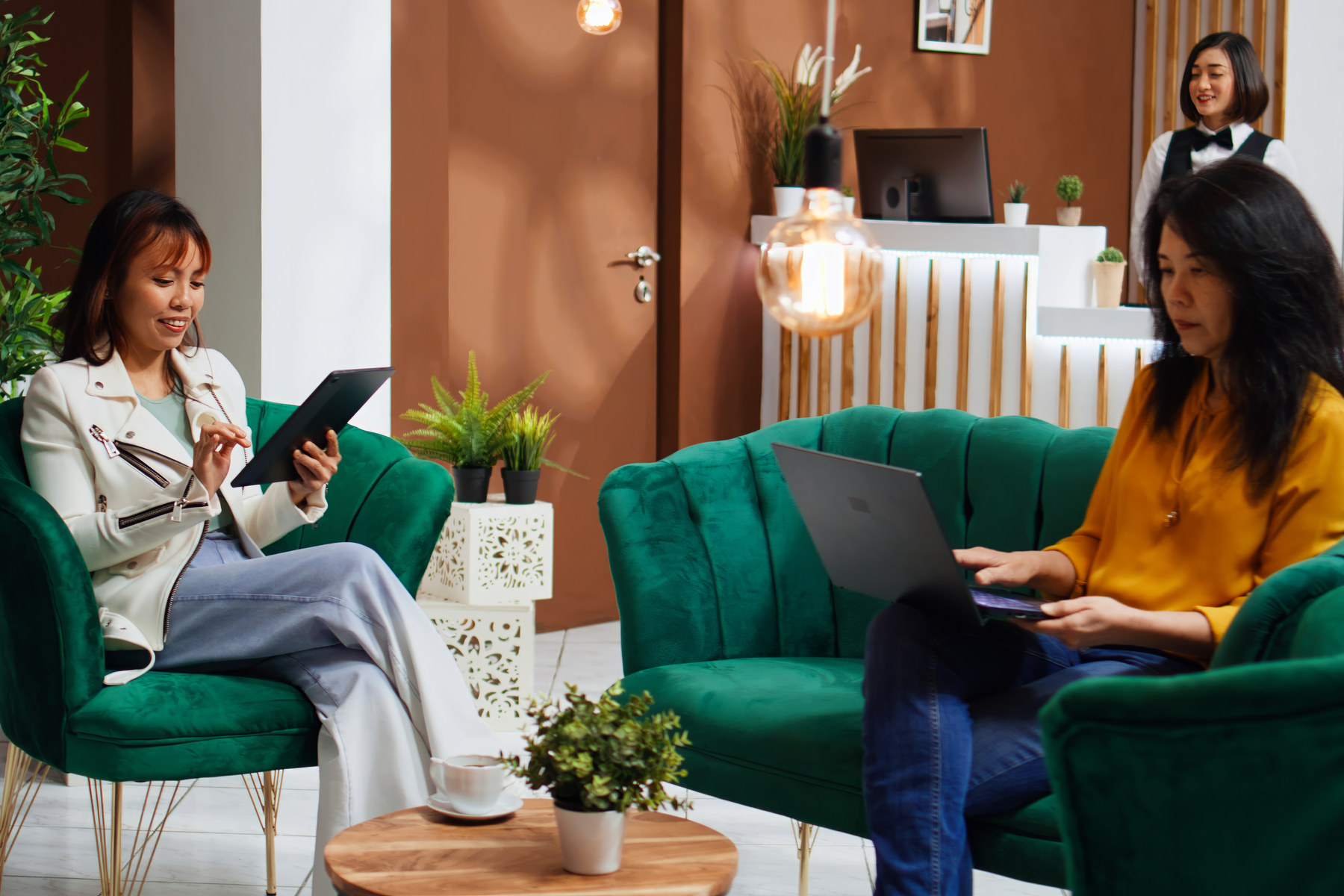 Two people sitting in armchairs in a terracotta-walled lounge with a low wooden table, potted plants and a glowing pendant lamp.