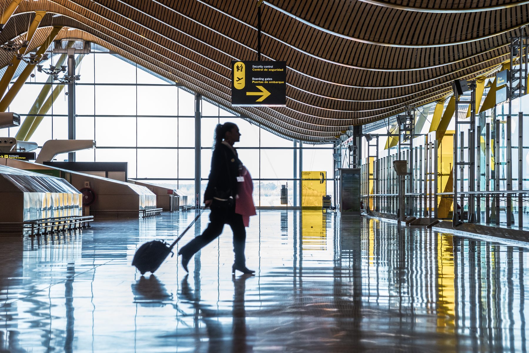 A traveller with a wheeled suitcase silhouetted against the glass wall of an airport terminal.