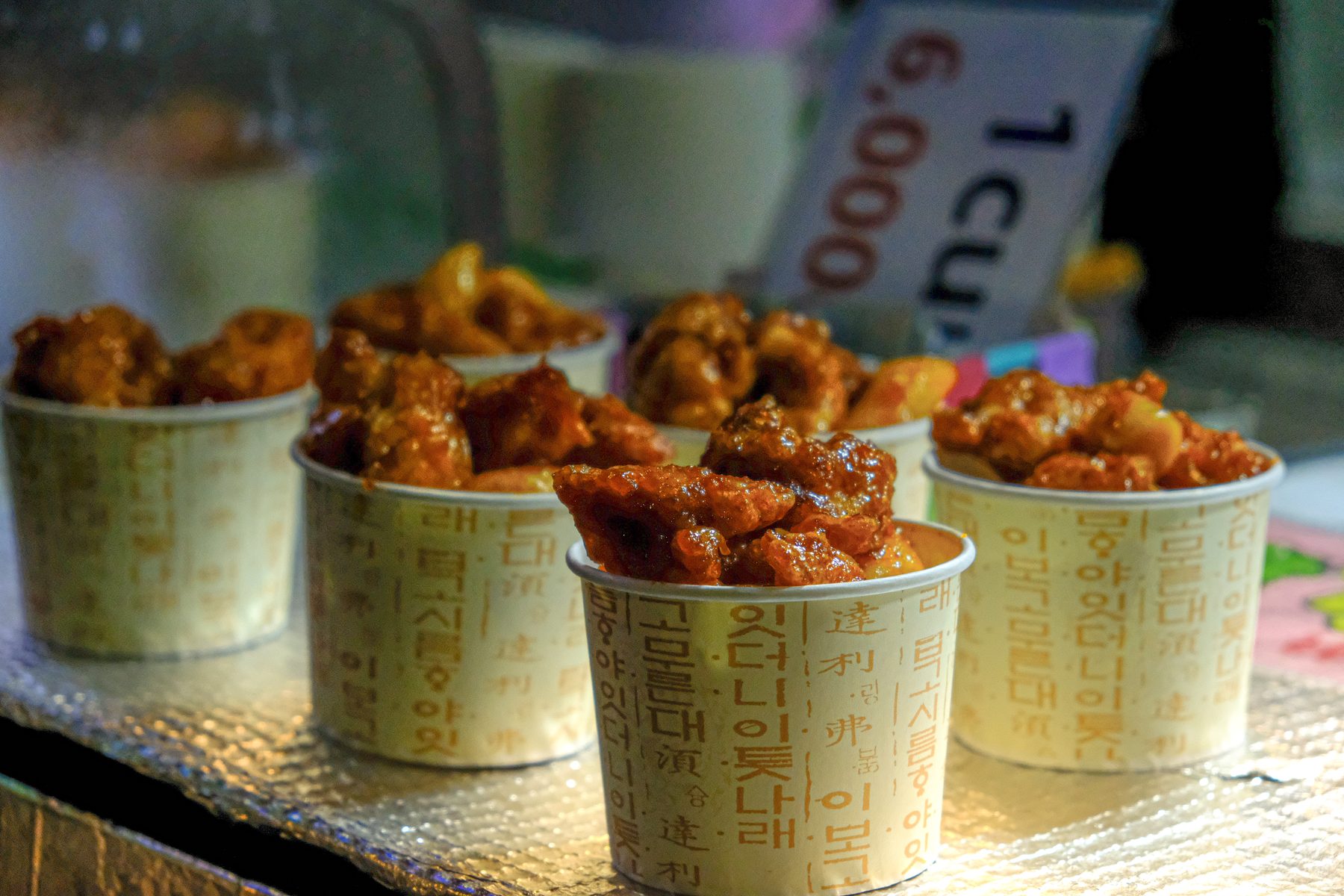 Paper cups of glazed Korean street-food chicken on a lit night-market counter in Seoul.